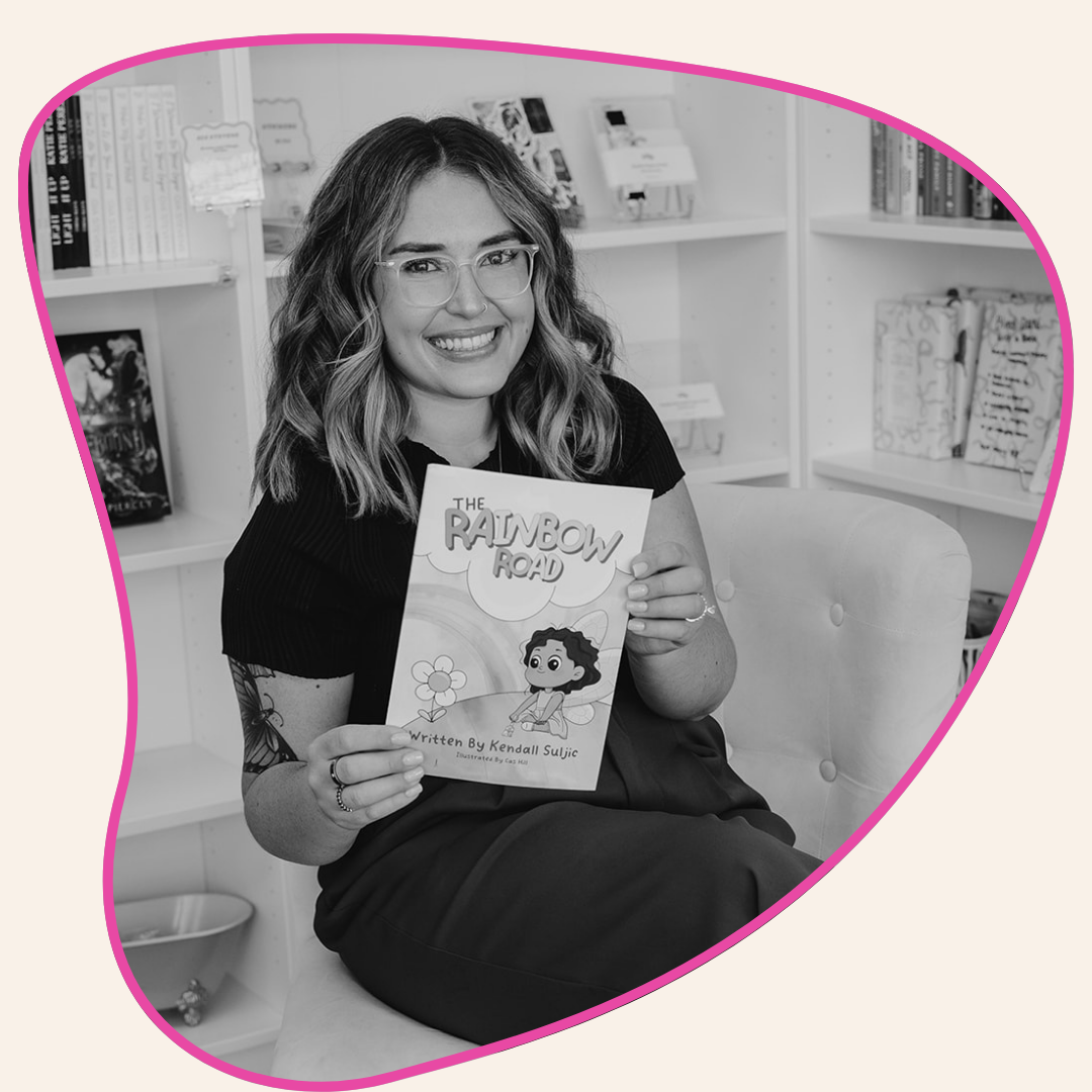 Woman holding a book titled 'The Rainbow Road' in a room with shelves.